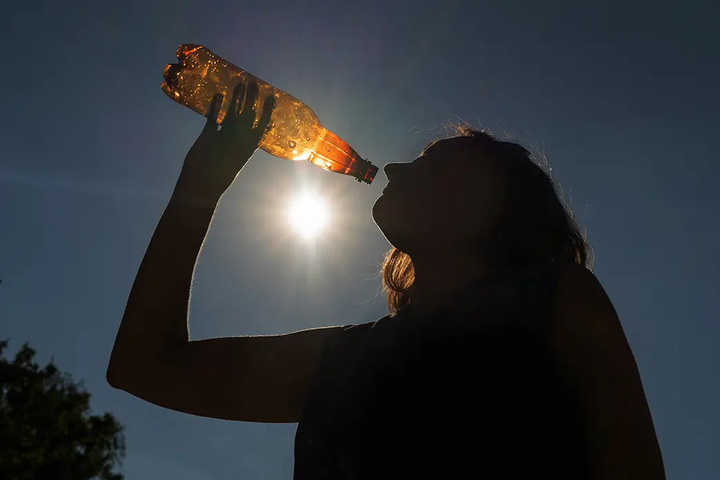 Trinken Sie an heißen Tagen mehr als sonst – vor allem, wenn Sie sich körperlich anstrengen Unter der grellen Sonne trinkt eine Frau Wasser aus einer Flasche