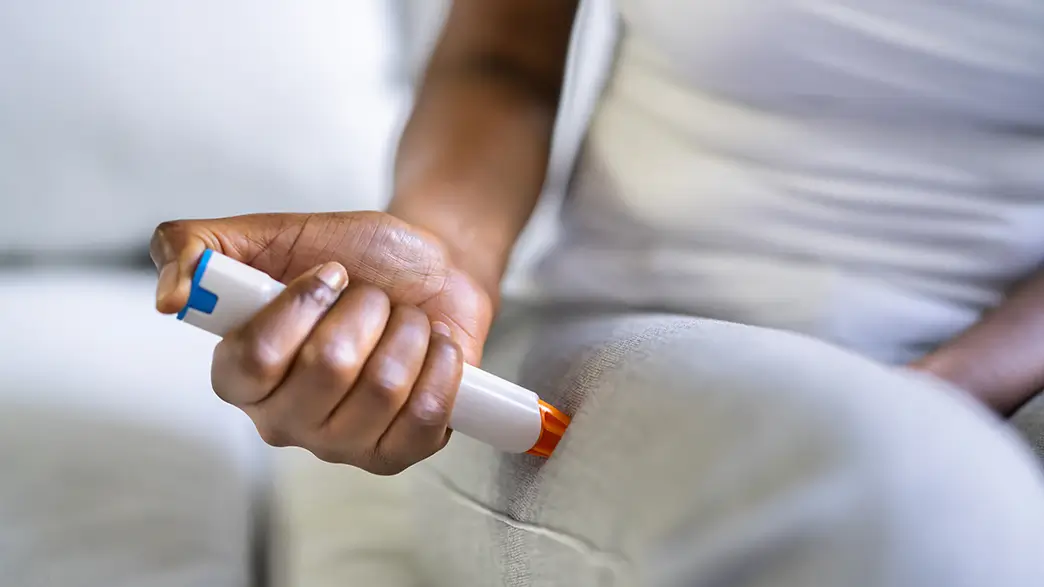 Das Notfallmedikament Epinephrin lässt sich im Falle eines allergischen Schocks direkt in den Oberschenkel injizieren Eine Frau in weißer Kleidung demonstriert den Einsatz eines Epinephrin-Injektors, der im Falle eines allergischen Schocks Adrenalin direkt in den Oberschenkelmuskel spritzt